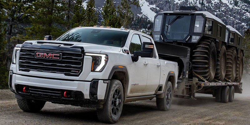 White 2026 GMC Sierra 2500HD towing a heavily-loaded off-road vehicle trailer on a countryside dirt road.